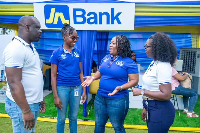 Four people chat at an outdoor Jn Bank booth under a blue-yellow canopy, smiling and engaging.