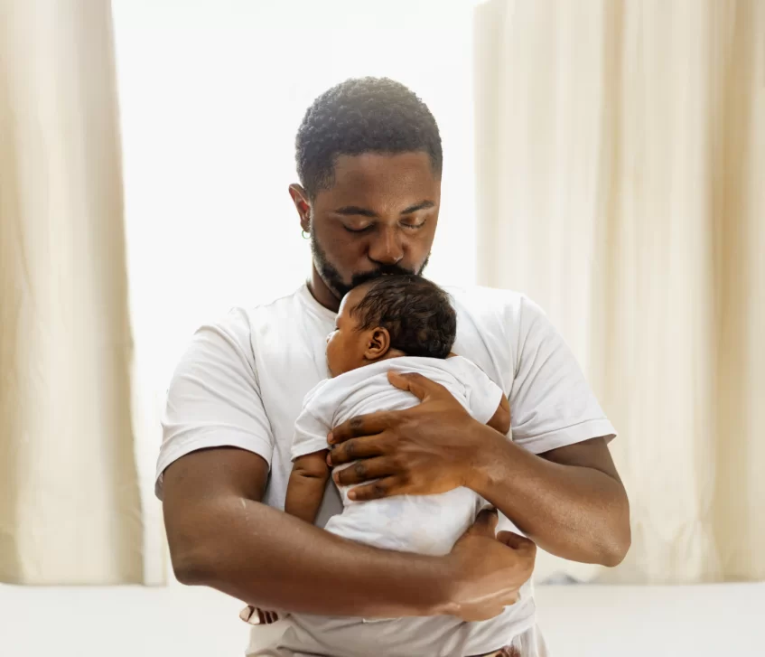 Father gently holding and kissing a newborn baby close to his chest indoors by light curtains.